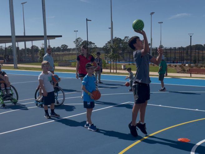 Children playing basketball on an outdoor court; one boy is jumping to shoot a green ball while others, including a child in a wheelchair, watch and wait their turn. It’s a sunny day.