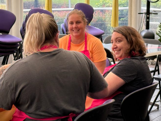 Three women wearing aprons sit together at a table, smiling and chatting in a brightly lit room with purple chairs stacked in the background.