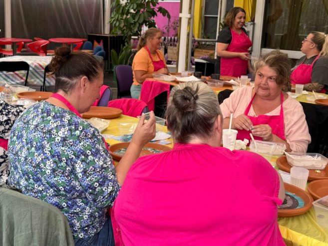 A group of women wearing pink aprons sit around a table, smiling and working on crafts or art projects together in a bright, colorful room.