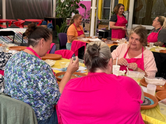 A group of women wearing pink aprons sit around a table, smiling and working on crafts or art projects together in a bright, colorful room.