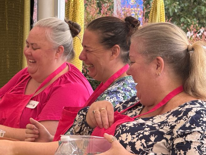 Three women wearing red aprons sit side by side, laughing together. They have their hair tied back and appear to be enjoying a group activity, possibly cooking or baking, with a water bottle and bowl in front of them.