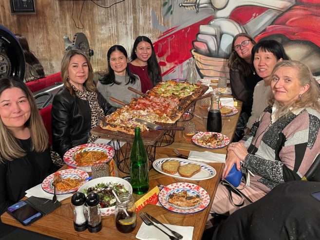 Seven women sit around a table in a restaurant, smiling at the camera. The table is filled with large pizzas, plates of pasta, salads, bread, and drinks. A colorful mural is visible on the wall behind them.