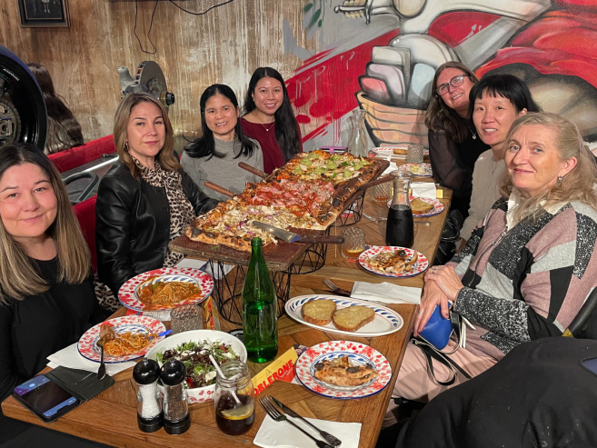 Seven women sit around a table in a restaurant, smiling at the camera. The table is filled with large pizzas, plates of pasta, salads, bread, and drinks. A colorful mural is visible on the wall behind them.