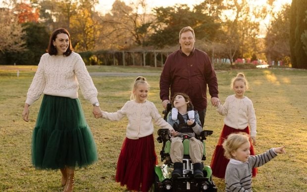 A family of six poses outdoors at sunset. The mother and three daughters wear cream sweaters and red or green skirts; the father stands behind a son in a wheelchair. A young boy runs ahead, and they all appear happy and relaxed.