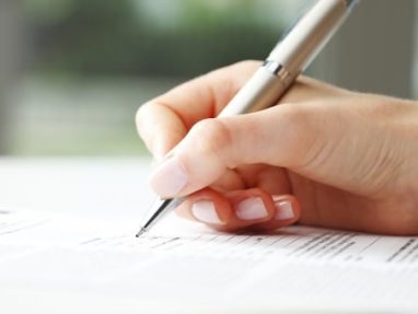 A close-up of a hand holding a silver pen and filling out a form or document on a white surface. The person has light skin and manicured nails. The background is blurred.