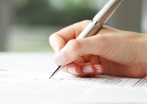 A close-up of a hand holding a silver pen and filling out a form or document on a white surface. The person has light skin and manicured nails. The background is blurred.