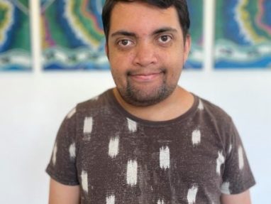 A man with short dark hair and a beard stands indoors, smiling at the camera. He is wearing a brown shirt with white patterns. Colorful abstract art is visible on the wall behind him.