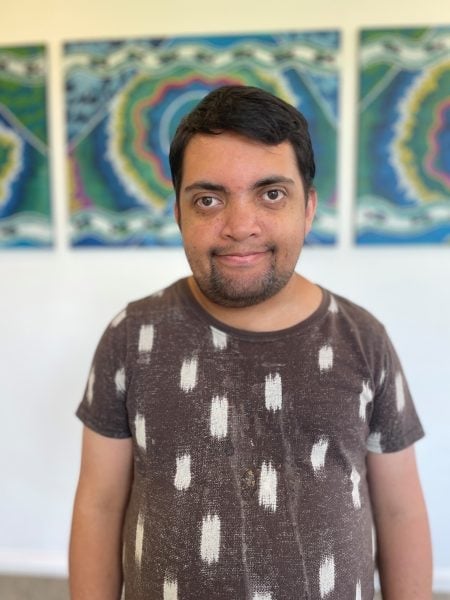 A man with short dark hair and a beard stands indoors, smiling at the camera. He is wearing a brown shirt with white patterns. Colorful abstract art is visible on the wall behind him.