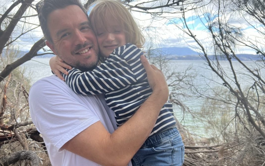 A man in a white t-shirt hugs a young child in a striped shirt. They are smiling, standing outdoors near a lake or sea, with trees and mountains in the background.