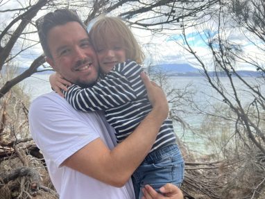 A man in a white t-shirt hugs a young child in a striped shirt. They are smiling, standing outdoors near a lake or sea, with trees and mountains in the background.