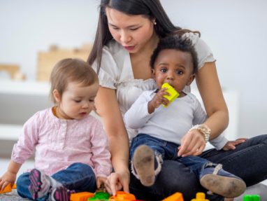 A woman sits on the floor with two toddlers, helping them play with colorful plastic building blocks. One child holds a yellow block in their mouth while the other child looks down, focused on the toys.