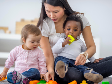 A woman sits on the floor with two toddlers, helping them play with colorful plastic building blocks. One child holds a yellow block in their mouth while the other child looks down, focused on the toys.