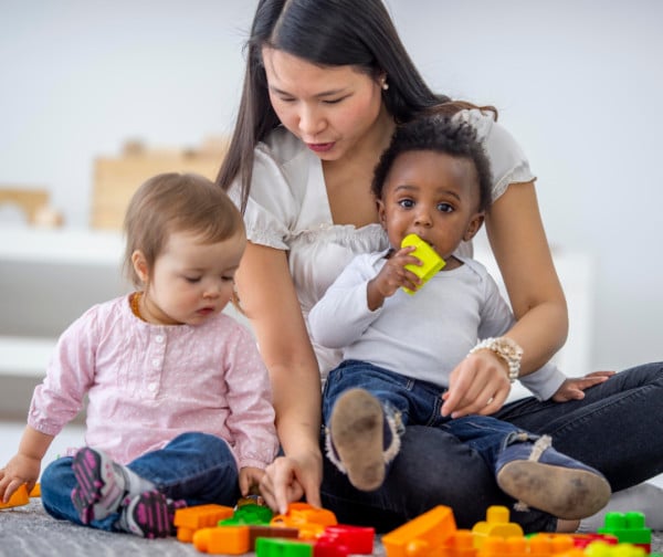 A woman sits on the floor with two toddlers, helping them play with colorful plastic building blocks. One child holds a yellow block in their mouth while the other child looks down, focused on the toys.