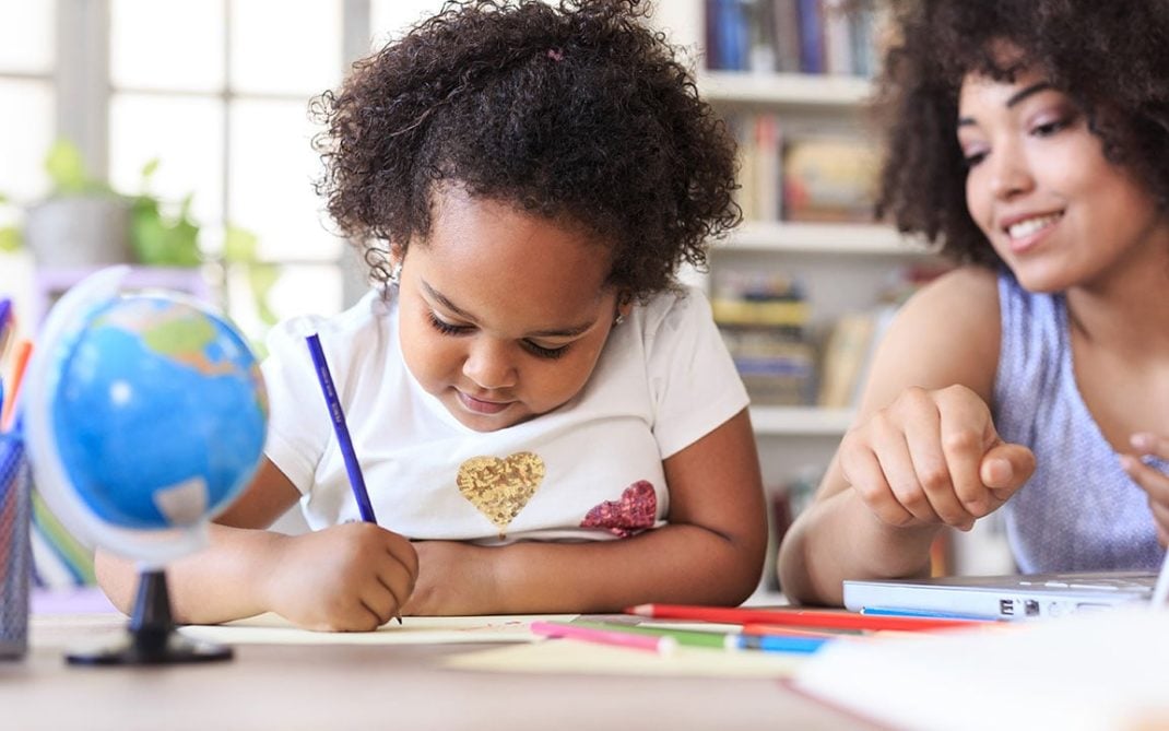 A young girl draws with a pencil at a table while an adult woman beside her smiles and points at something. Colored pencils, a small globe, and books are visible in the background.
