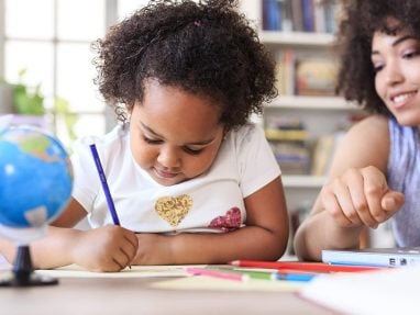 A young girl draws with a pencil at a table while an adult woman beside her smiles and points at something. Colored pencils, a small globe, and books are visible in the background.