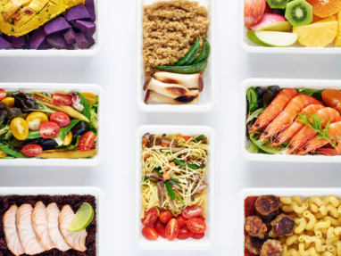 A variety of healthy meal prep containers with foods including grilled chicken, shrimp, pasta, salads, vegetables, fruits, noodles, and quinoa, neatly arranged on a white background.