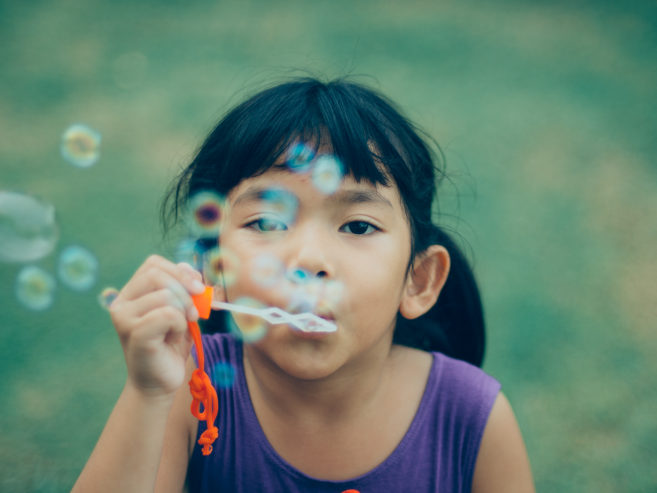 A young girl with dark hair and a purple top blows soap bubbles outdoors, with blurred green grass in the background.