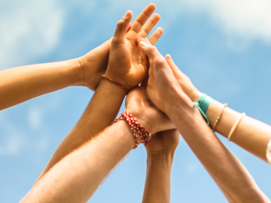 Several hands reaching up and touching together against a blue sky, symbolizing unity, teamwork, and support. Some hands wear colorful bracelets.