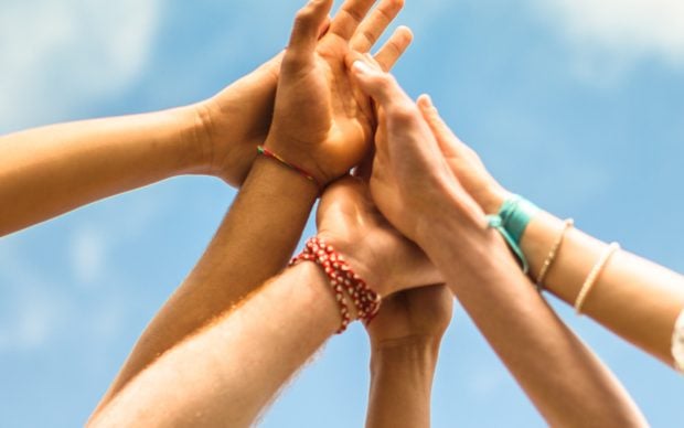 Several hands reaching up and touching together against a blue sky, symbolizing unity, teamwork, and support. Some hands wear colorful bracelets.
