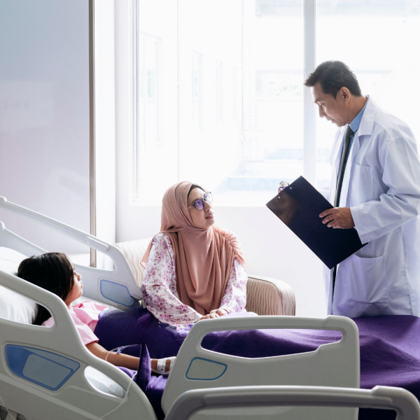 A doctor holding a clipboard stands beside a hospital bed, talking to a woman in a hijab and glasses, who is sitting next to a child lying under a purple blanket in the hospital room.