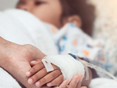 A child in a hospital bed with a bandaged hand and IV, holding an adults hand for comfort. The focus is on their hands, symbolizing care and support.