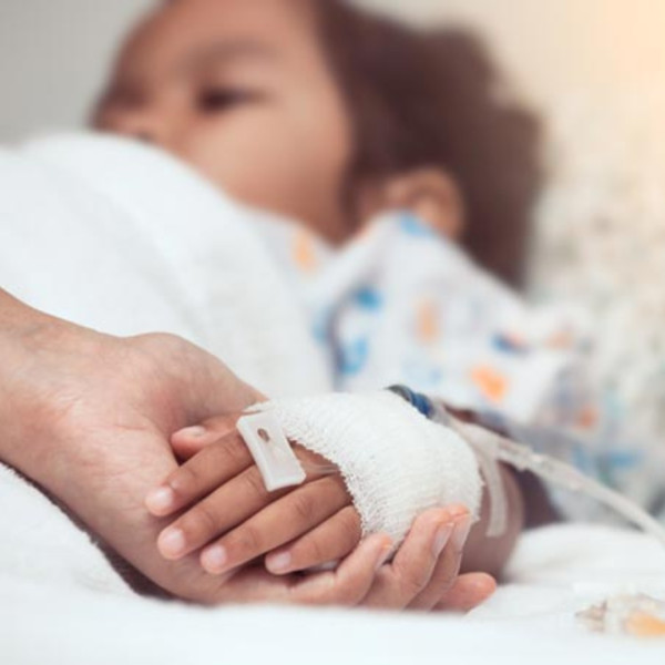 A child in a hospital bed with a bandaged hand and IV, holding an adults hand for comfort. The focus is on their hands, symbolizing care and support.