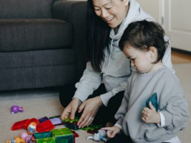 An adult and a young child sit on the carpet, smiling and playing with colorful magnetic building tiles and toys in a living room.