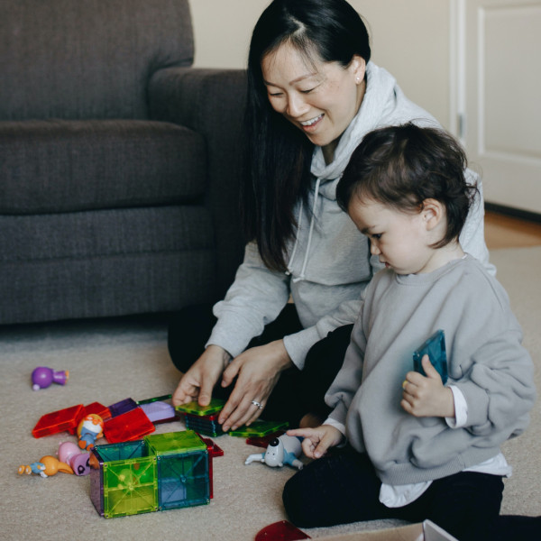 An adult and a young child sit on the carpet, smiling and playing with colorful magnetic building tiles and toys in a living room.