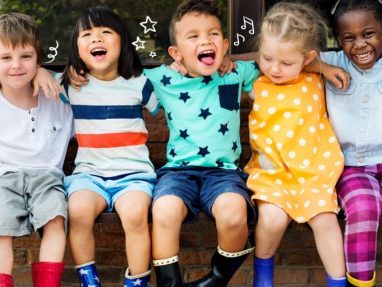 Five young children sit close together on a bench outdoors, smiling and laughing. They wear colorful outfits and rain boots, with doodles of stars and music notes drawn above their heads.