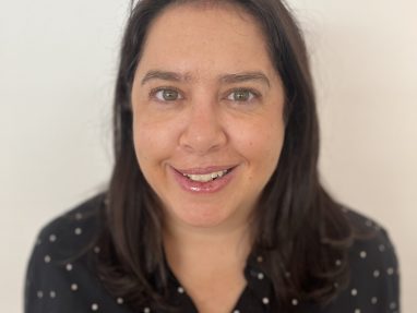 A woman with straight, dark brown hair and green eyes, wearing a black blouse with white polka dots, smiles at the camera against a plain white background.