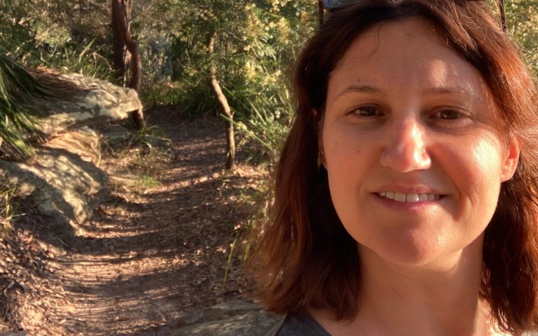 A woman with brown hair and sunglasses on her head smiles at the camera while standing on a sunlit, wooded hiking trail surrounded by trees and rocks.