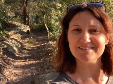A woman with brown hair and sunglasses on her head smiles at the camera while standing on a sunlit, wooded hiking trail surrounded by trees and rocks.