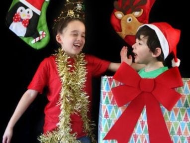 Two children celebrate Christmas; one wears a tinsel garland and tree hat, the other sits in a large gift box with a Santa hat. Festive stockings hang in the background against a black backdrop.
