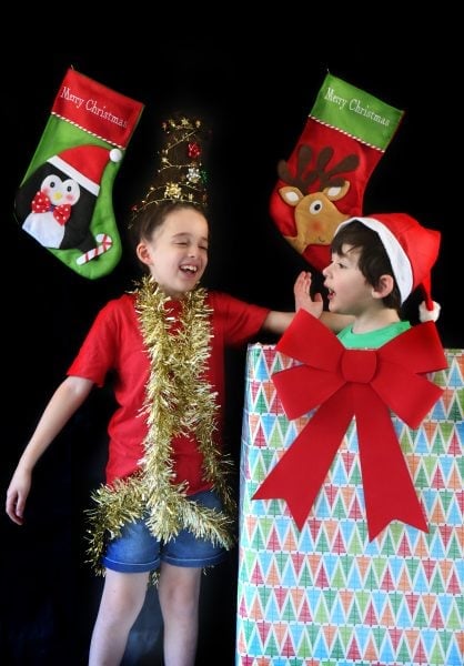 Two children celebrate Christmas; one wears a tinsel garland and tree hat, the other sits in a large gift box with a Santa hat. Festive stockings hang in the background against a black backdrop.