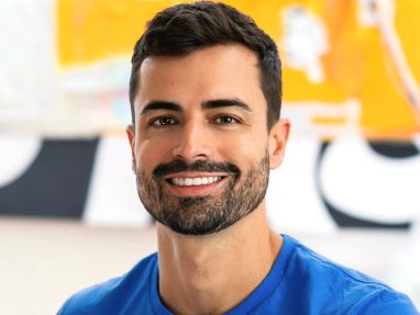 A smiling man with short dark hair and a beard, wearing a bright blue shirt, sits in front of a colorful abstract background.