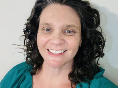 A woman with curly brown hair and blue eyes smiles at the camera. She is wearing a teal blouse and standing in front of a plain light-colored wall.