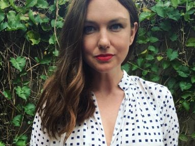 A woman with long brown hair stands in front of a leafy green background, wearing a white blouse with a blue square pattern and bright red lipstick, looking directly at the camera.