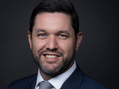 A man with short dark hair, a beard, and blue eyes is smiling and wearing a navy suit, white shirt, and patterned tie against a dark background.