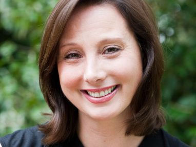 A woman with shoulder-length brown hair smiles at the camera. She is wearing a sleeveless black top and a necklace with a circular pendant. Green foliage is blurred in the background.