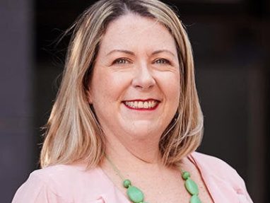 A woman with shoulder-length blonde hair smiles at the camera. She is wearing a light pink blouse and a chunky green bead necklace, standing in front of a dark, blurred background.