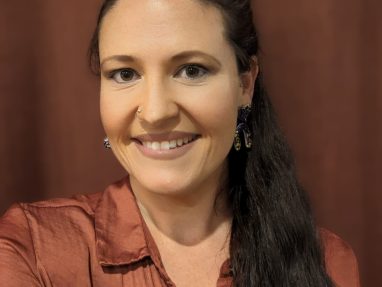 A woman with long dark hair in a ponytail, wearing a rust-colored blouse and dangling earrings, smiles at the camera against a brown background.