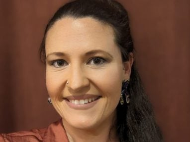 A woman with long dark hair in a ponytail, wearing a rust-colored blouse and dangling earrings, smiles at the camera against a brown background.