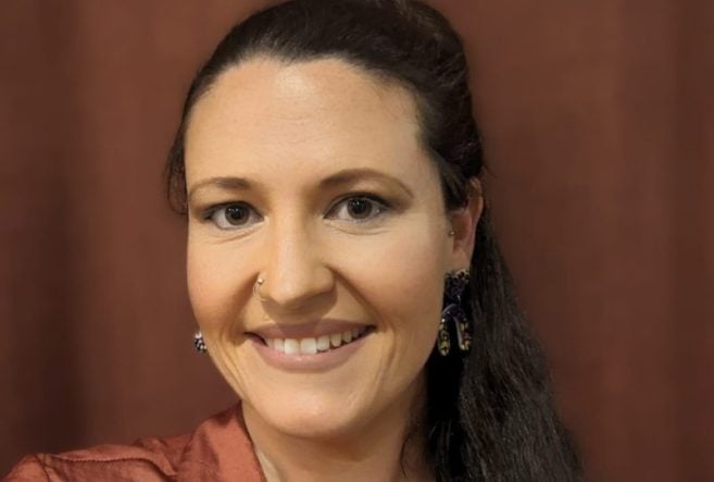A woman with long dark hair in a ponytail, wearing a rust-colored blouse and dangling earrings, smiles at the camera against a brown background.