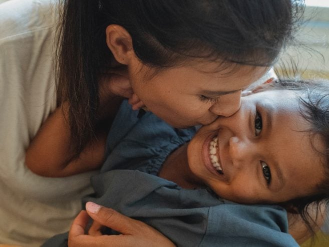A woman lovingly kisses a smiling young child on the cheek as they embrace at a table. The child is looking at the camera, beaming with joy, and both appear happy and affectionate.