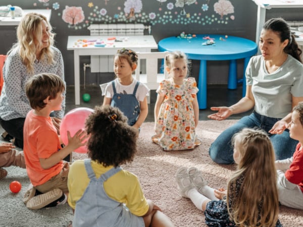 A group of young children sit in a circle on a classroom rug, listening to two adult women. The room is decorated with colorful wall art and children’s tables in the background.