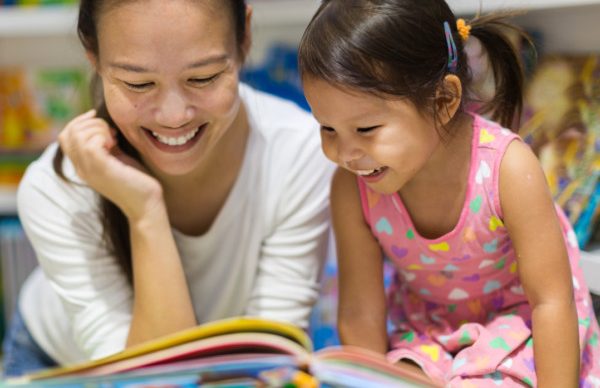 An adult and a young girl sit close together, smiling and reading a colorful childrens book in a brightly lit room filled with books.