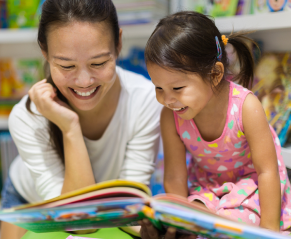 An adult and a young girl sit close together, smiling and reading a colorful childrens book in a brightly lit room filled with books.