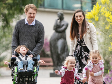 A smiling family enjoys a walk outdoors. A man pushes a boy in a wheelchair while a woman walks beside him. In front, two young girls hold hands and run together, surrounded by greenery and flowers.