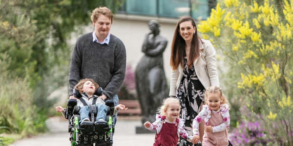 A smiling family enjoys a walk outdoors. A man pushes a boy in a wheelchair while a woman walks beside him. In front, two young girls hold hands and run together, surrounded by greenery and flowers.