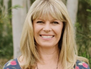 A smiling woman with long blonde hair and bangs stands outdoors in front of trees, wearing a patterned top and a silver necklace.